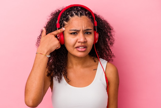 Giovane donna afroamericana che ascolta la musica con le cuffie isolate sulla parete rosa che mostra un gesto di delusione con l'indice