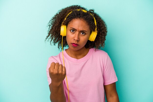 Giovane donna afroamericana che ascolta musica isolata su fondo blu che mostra pugno alla macchina fotografica, espressione facciale aggressiva.