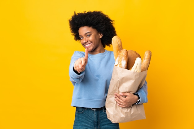 Giovane donna afroamericana che compra qualcosa di pane sulla parete gialla che mostra e che solleva un dito