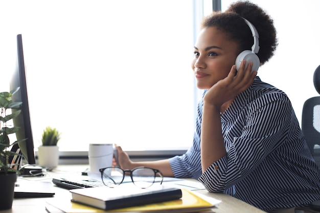 Giovane donna afroamericana che ha una pausa e ascolta musica in cuffia seduta sul posto di lavoro.