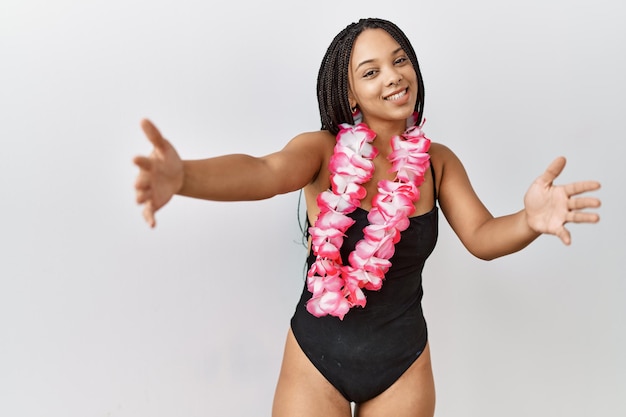 Giovane donna afroamericana che indossa costume da bagno e lei hawaiana guardando la telecamera sorridendo a braccia aperte per l'abbraccio espressione allegra che abbraccia la felicità