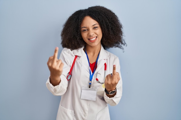 Giovane donna afroamericana che indossa l'uniforme del medico e lo stetoscopio che mostra il dito medio facendoti fottere cattiva espressione, provocazione e atteggiamento maleducato. urlando eccitato