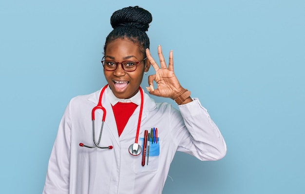 Giovane donna afroamericana che indossa l'uniforme del medico e lo stetoscopio che sorride positivo facendo segno ok con la mano e le dita espressione di successo