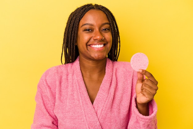 Giovane donna afroamericana che indossa un accappatoio in possesso di un disco per il trucco isolato su sfondo giallo che ride e si diverte.