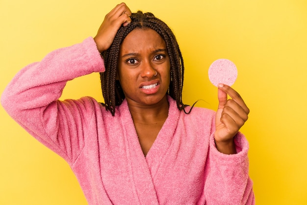 Giovane donna afroamericana che indossa un accappatoio in possesso di un disco per il trucco isolato su sfondo giallo scioccata, ha ricordato un incontro importante.