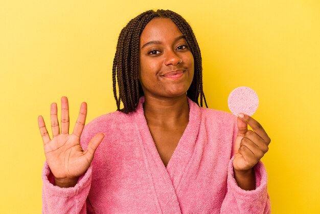 Giovane donna afroamericana che indossa un accappatoio in possesso di un disco per il trucco isolato su sfondo giallo sorridente allegro che mostra il numero cinque con le dita.