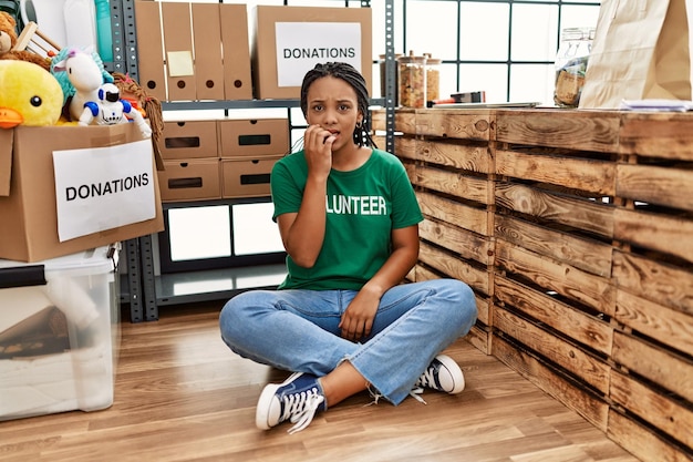 Giovane donna afroamericana che indossa una maglietta volontaria allo stand delle donazioni che sembra stressata e nervosa con le mani sulla bocca che mordono il problema dell'ansia delle unghie