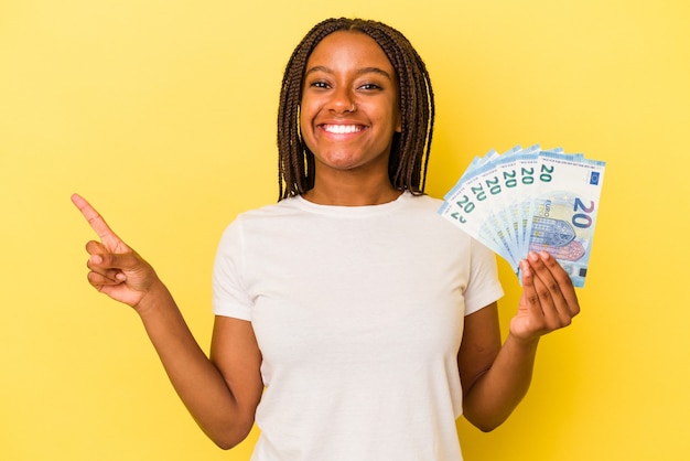 Giovane donna afroamericana che tiene banconote isolate su sfondo giallo sorridendo e indicando da parte, mostrando qualcosa in uno spazio vuoto.