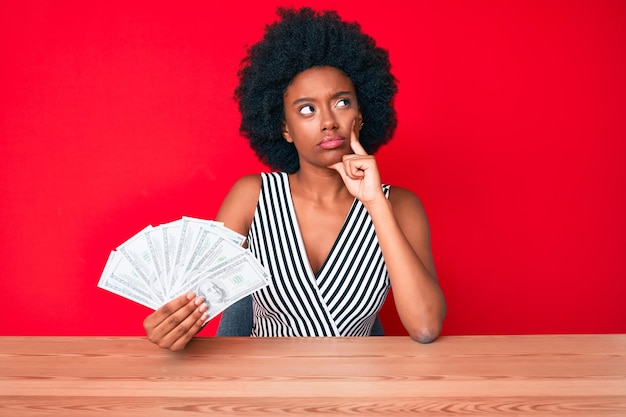 Giovane donna afroamericana che tiene dollari faccia seria pensando alla domanda con la mano sul mento premurosa sull'idea confusa