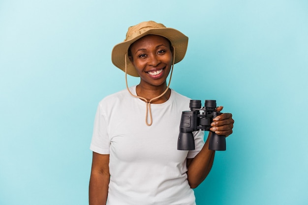 Giovane donna afroamericana che tiene il binocolo isolato su sfondo blu felice, sorridente e allegro.