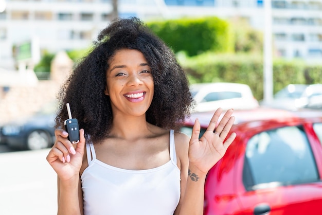 Giovane donna afroamericana che tiene le chiavi della macchina all'aperto salutando con la mano con un'espressione felice
