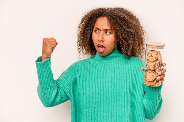 Giovane donna afroamericana che tiene un barattolo di biscotti isolato su sfondo bianco alzando il pugno dopo un concetto di vincitore della vittoria