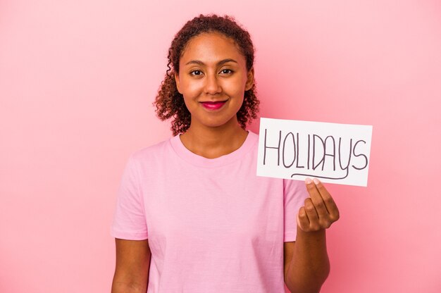 Giovane donna afroamericana che tiene un cartello di vacanze isolato su sfondo rosa