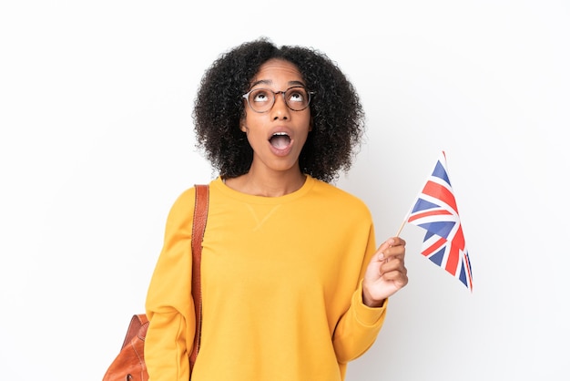 Giovane donna afroamericana che tiene una bandiera del Regno Unito isolata su fondo bianco che guarda in su e con espressione sorpresa