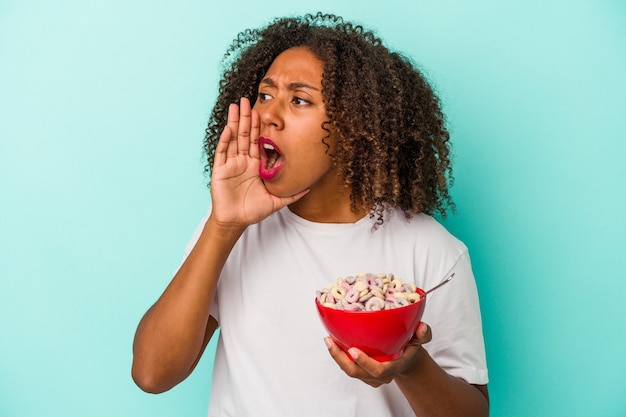 Giovane donna afroamericana che tiene una ciotola di cereali isolata su fondo blu che grida e che tiene il palmo vicino alla bocca aperta.