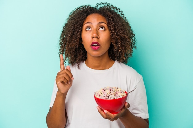 Giovane donna afroamericana che tiene una ciotola di cereali isolata su fondo blu che indica al rialzo con la bocca aperta.