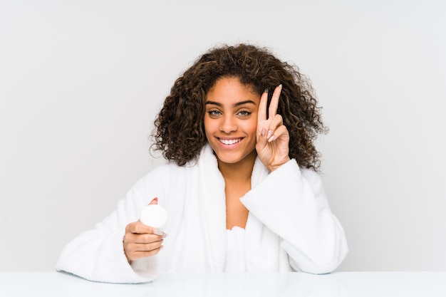 Giovane donna afroamericana che tiene una crema idratante che mostra il segno di vittoria e che sorride ampiamente.