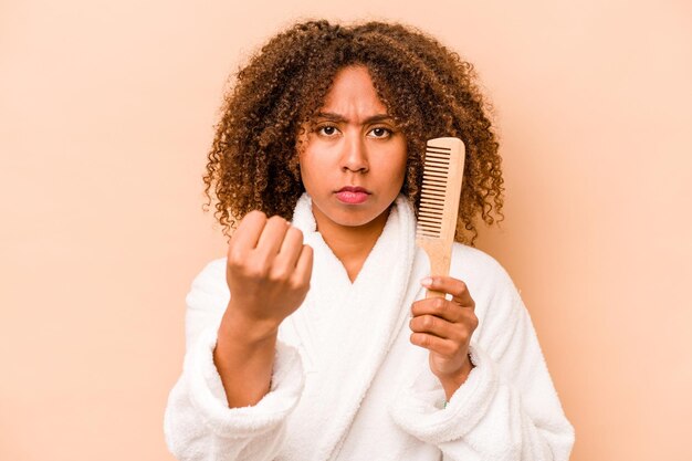 Giovane donna afroamericana che tiene una spazzola per capelli isolata su sfondo beige che mostra il pugno all'espressione facciale aggressiva della fotocamera