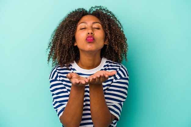 Giovane donna afroamericana con capelli ricci isolati su sfondo blu piegando le labbra e tenendo le palme per inviare un bacio d'aria.