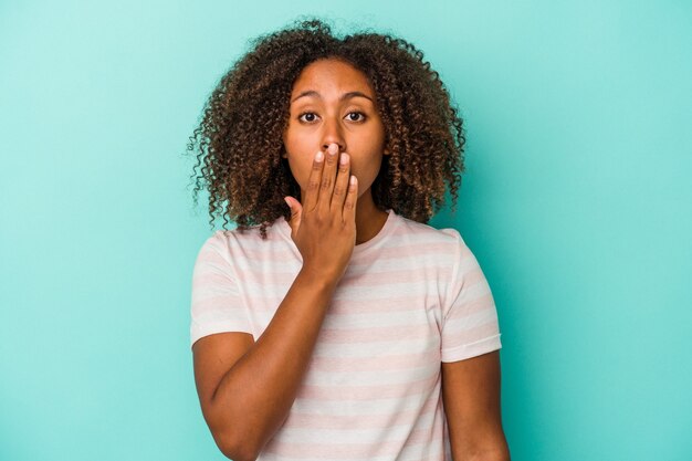 Giovane donna afroamericana con i capelli ricci isolata su sfondo blu scioccata, coprendo la bocca con le mani, ansiosa di scoprire qualcosa di nuovo.