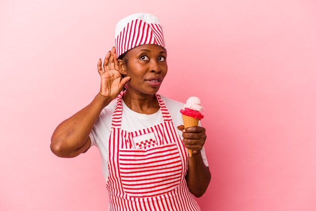 Giovane donna afroamericana del gelatiere che tiene il gelato isolato su sfondo rosa cercando di ascoltare un pettegolezzo.