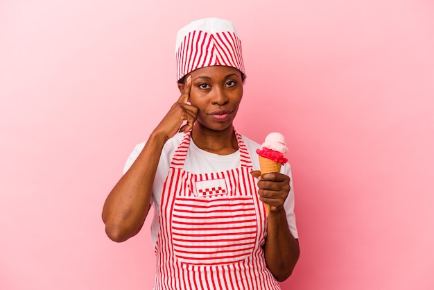 Giovane donna afroamericana del gelatiere che tiene il gelato isolato su sfondo rosa che punta il tempio con il dito, pensando, concentrato su un compito.