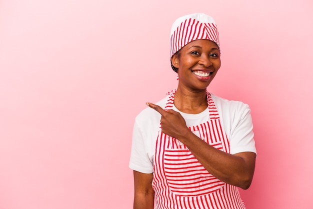 Giovane donna afroamericana del gelatiere isolata su fondo rosa che sorride e che indica da parte, mostrando qualcosa allo spazio vuoto.