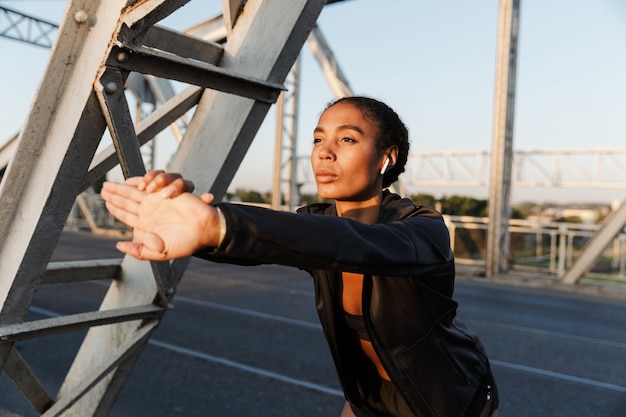 giovane donna afroamericana in abbigliamento sportivo usando auricolari e allungando le braccia mentre si allena sul vecchio ponte