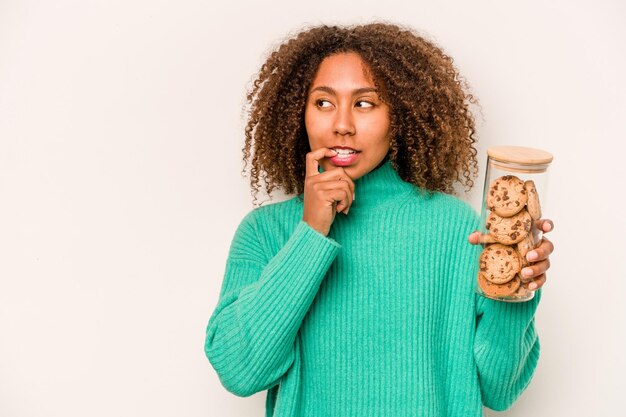 Giovane donna afroamericana in possesso di un vasetto di biscotti isolato su sfondo bianco rilassato pensando a qualcosa guardando uno spazio di copia