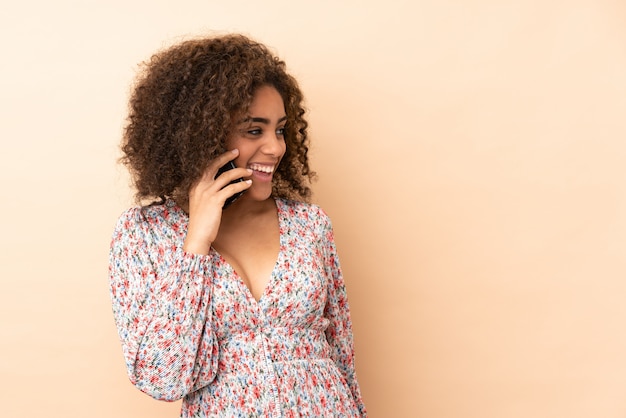 Giovane donna afroamericana isolata su beige che mantiene una conversazione con il telefono cellulare con qualcuno