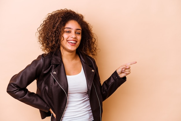 Giovane donna afroamericana isolata su fondo beige che sorride allegramente indicando con l'indice via.