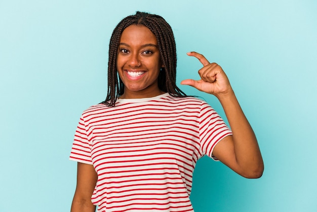 Giovane donna afroamericana isolata su fondo blu che tiene qualcosa di piccolo con l'indice, sorridente e fiducioso.