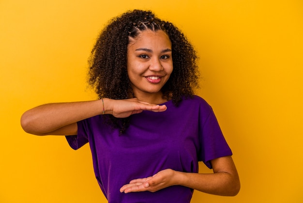 Giovane donna afroamericana isolata su fondo giallo che tiene qualcosa con entrambe le mani, presentazione del prodotto.