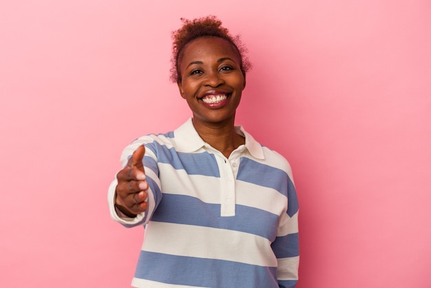Giovane donna afroamericana isolata su fondo rosa che allunga la mano alla macchina fotografica nel gesto di saluto.