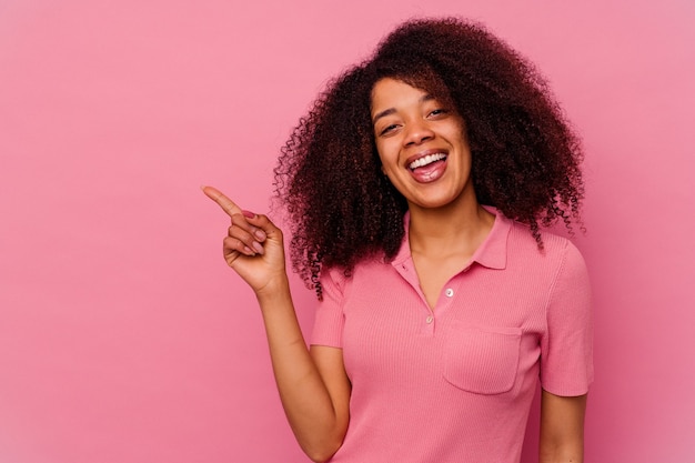 Giovane donna afroamericana isolata su fondo rosa che sorride allegramente indicando con l'indice via.