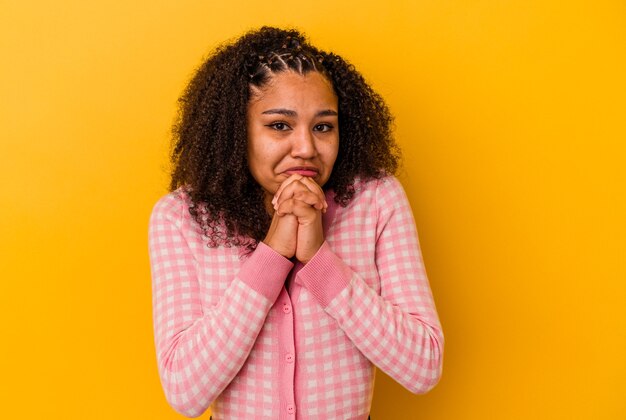 Giovane donna afroamericana isolata su priorità bassa gialla che prega per fortuna, bocca stupita e di apertura che osserva alla parte anteriore.