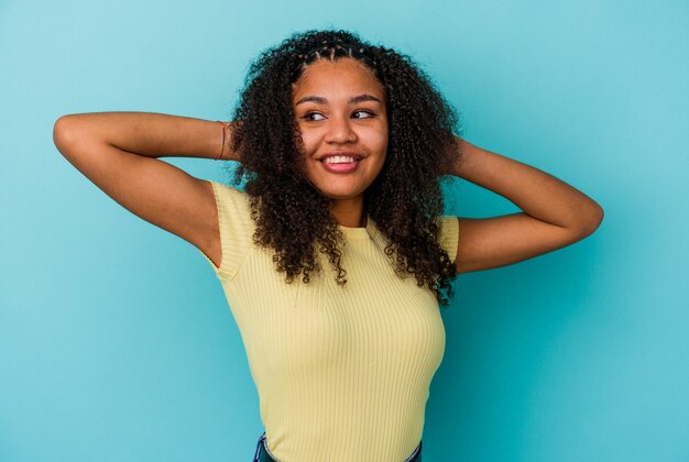 Giovane donna afroamericana isolata su sfondo blu fiduciosa, con le mani dietro la testa.