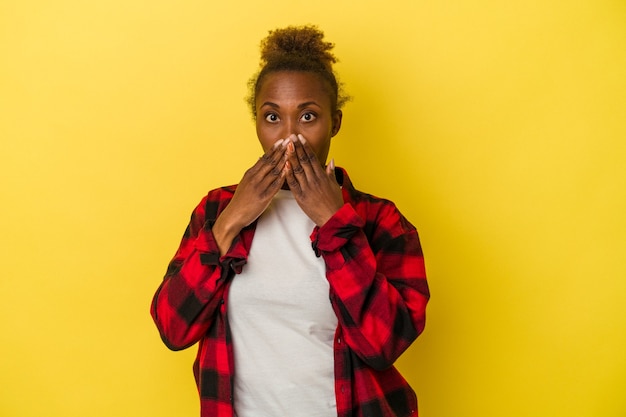 Giovane donna afroamericana isolata su sfondo giallo scioccato che copre la bocca con le mani.