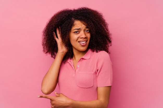 Giovane donna afroamericana isolata su sfondo rosa, cercando di ascoltare un pettegolezzo.