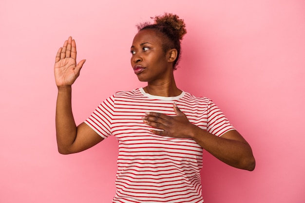 Giovane donna afroamericana isolata su sfondo rosa prestando giuramento, mettendo la mano sul petto.