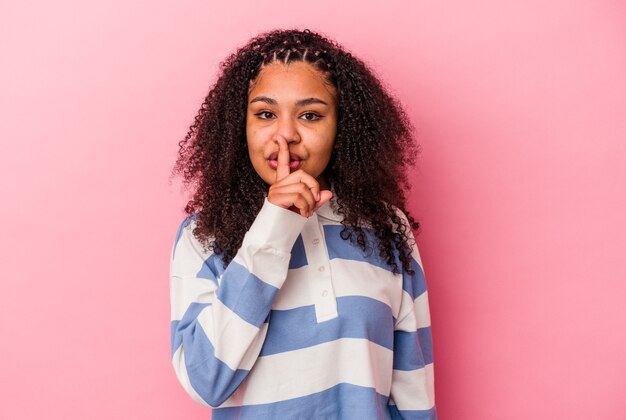 Giovane donna afroamericana isolata sul muro rosa mantenendo un segreto o chiedendo silenzio.