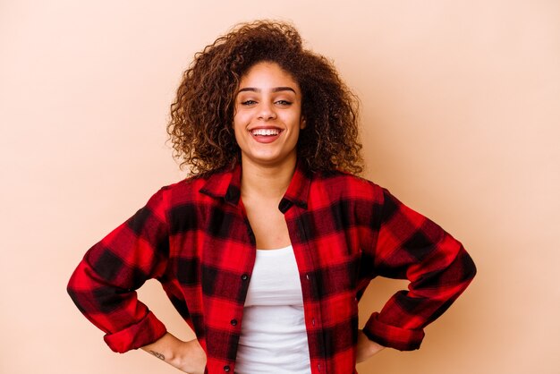 Giovane donna afroamericana isolata sulla parete beige fiduciosa mantenendo le mani sui fianchi.