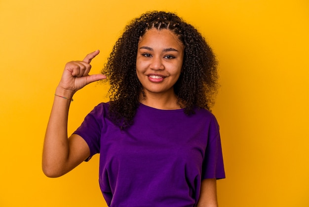 Giovane donna afroamericana isolata sulla parete gialla che tiene qualcosa di poco con gli indici, sorridente e sicura