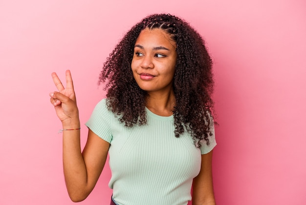 Giovane donna afroamericana isolata sulla parete rosa gioiosa e spensierata che mostra un simbolo di pace con le dita.