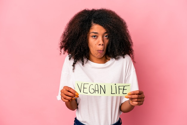 Giovane donna afroamericana riccia che tiene un cartello di vita vegano alza le spalle e apre gli occhi confusi.