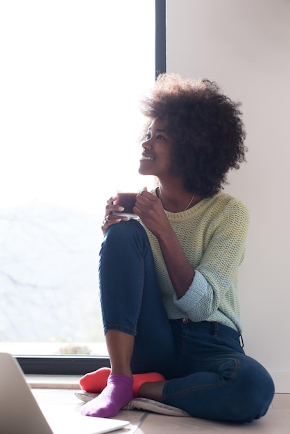 Giovane donna afroamericana sorridente seduta sul pavimento vicino alla finestra luminosa mentre guarda il computer portatile aperto e tiene la tazza a casa