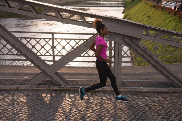 Giovane donna afroamericana sportiva che corre sul marciapiede attraverso il ponte al mattino presto a fare jogging con la scena dell'alba della città sullo sfondo