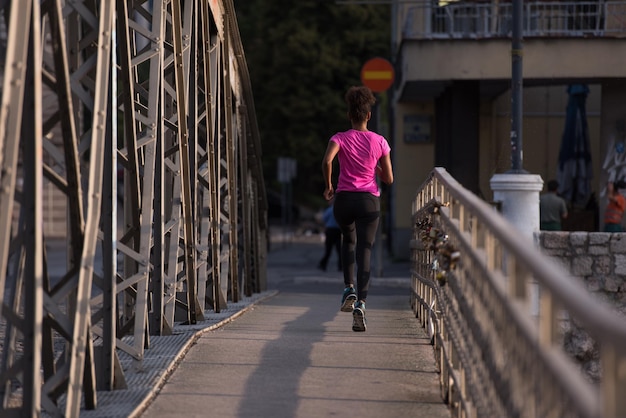 Giovane donna afroamericana sportiva che corre sul marciapiede attraverso il ponte al mattino presto a fare jogging con la scena dell'alba della città sullo sfondo
