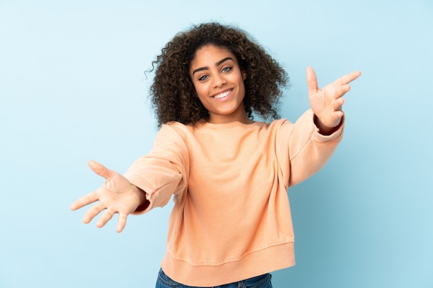 Giovane donna afroamericana sulla parete blu che presenta e che invita a venire con la mano