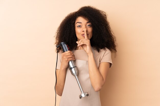 Giovane donna afroamericana utilizzando frullatore a immersione isolato su beige facendo gesto di silenzio